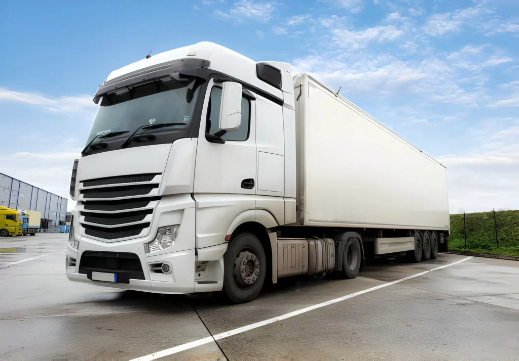A large white commercial truck is parked on a wet road under a cloudy sky, representing a trusted trucking company in UAE. The industrial setting evokes a sense of movement and logistics.