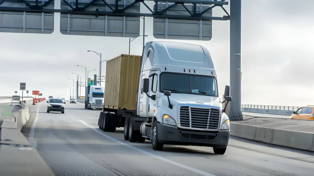 A white semi-truck with a brown container drives on a highway bridge, representing a trusted trucking company in UAE. The sky is overcast, and other vehicles are visible in the background.