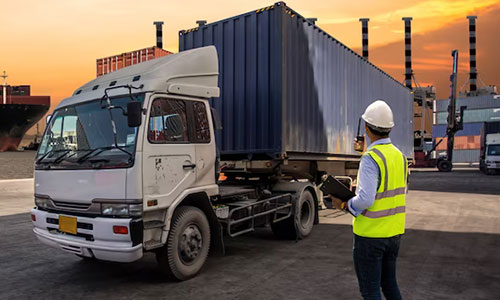 A worker in a safety vest and helmet uses a tablet while standing near a white truck with a cargo container at sunset, representing truck rental Dubai and logistics operations.
