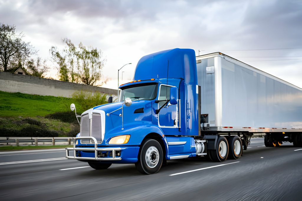 A blue semi-truck with a white trailer drives on a highway under a cloudy sky, surrounded by green grassy banks, representing heavy truck rental Dubai services and conveying a sense of motion and purpose. pickup truck delivery Dubai.