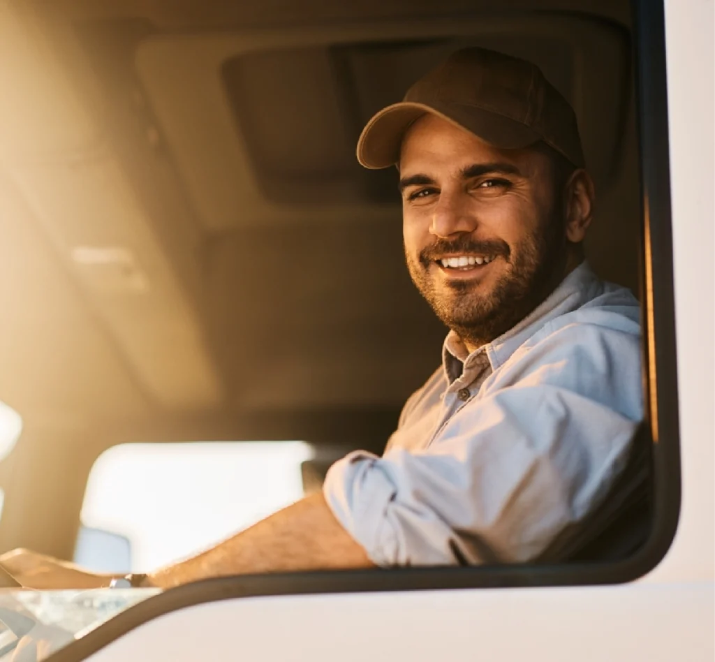 Smiling man in a baseball cap and blue shirt sitting in a vehicle's driver seat, illuminated by warm sunlight, representing a trusted trucking company in UAE and conveying a cheerful, relaxed mood.