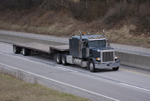 A blue semi-truck with a long flatbed trailer travels on an empty highway surrounded by grassy hills, creating a sense of motion and solitude.