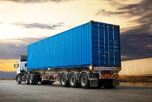 A large blue shipping container on a semi-trailer truck is parked on a road at sunset, representing heavy truck rental Dubai services. The dramatic sky in the background conveys a calm, expansive feel.