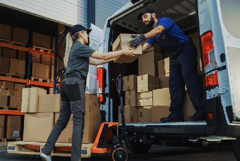 Two delivery workers load cardboard boxes into a van from a warehouse, representing pickup truck delivery Dubai services and conveying teamwork and efficiency in a logistics setting.