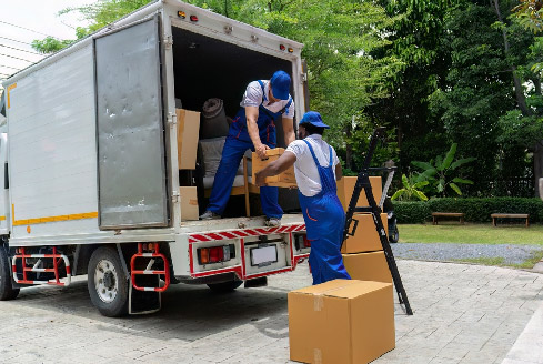 Two movers in blue uniforms load brown boxes into a truck parked on a street lined with trees, representing pickup truck delivery Dubai services. A ladder leans against the open truck.