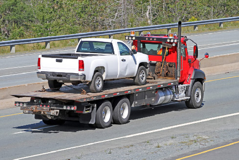 A red tow truck hauls a white pickup truck on a flatbed along a highway, representing trusted car recovery Dubai services. The road is bordered by greenery, conveying a sense of transit.