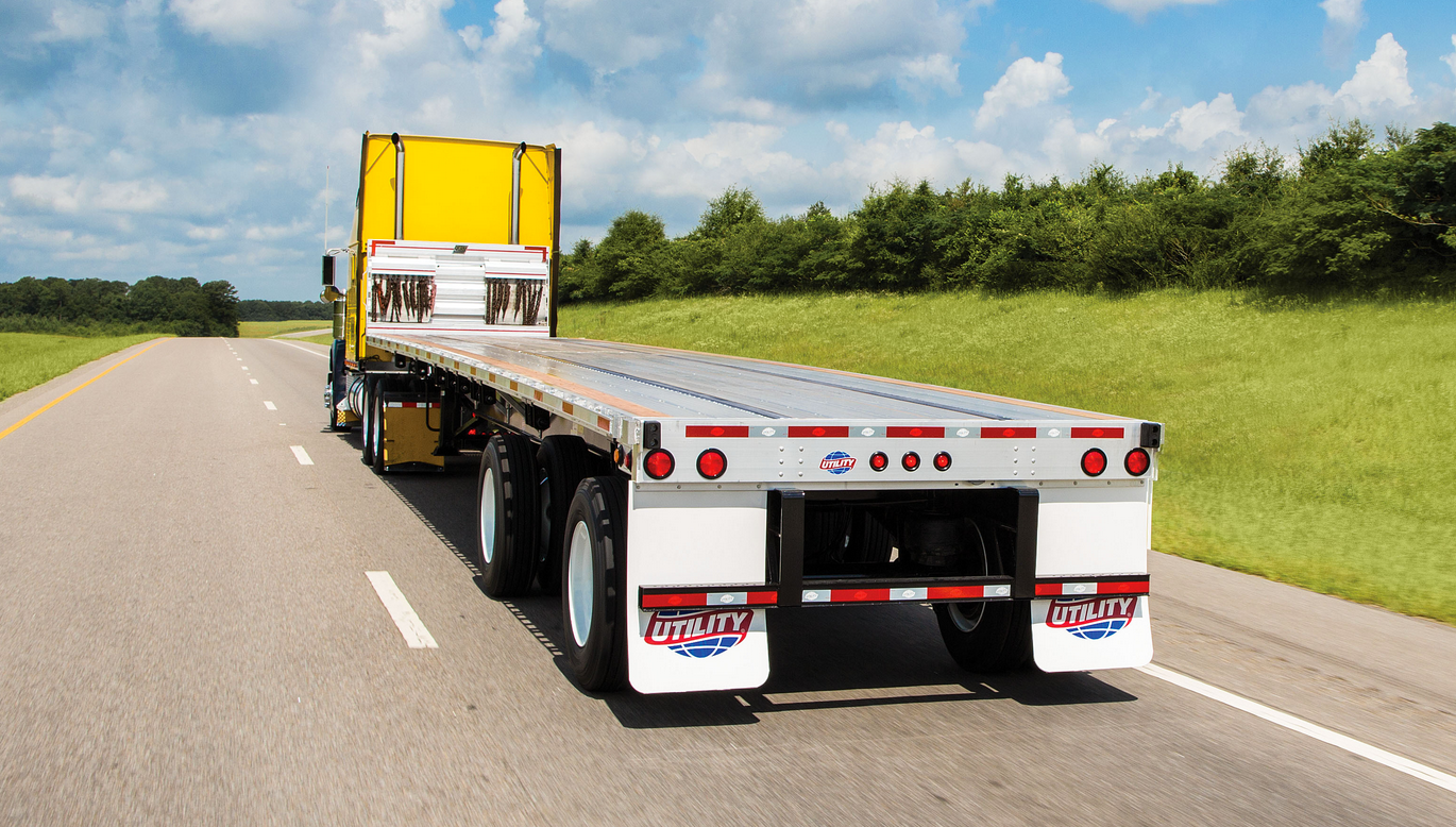 A flatbed truck with a yellow cab travels on a highway surrounded by green fields under a blue sky with clouds, representing flatbed truck rental Dubai services and conveying a sense of movement and freedom