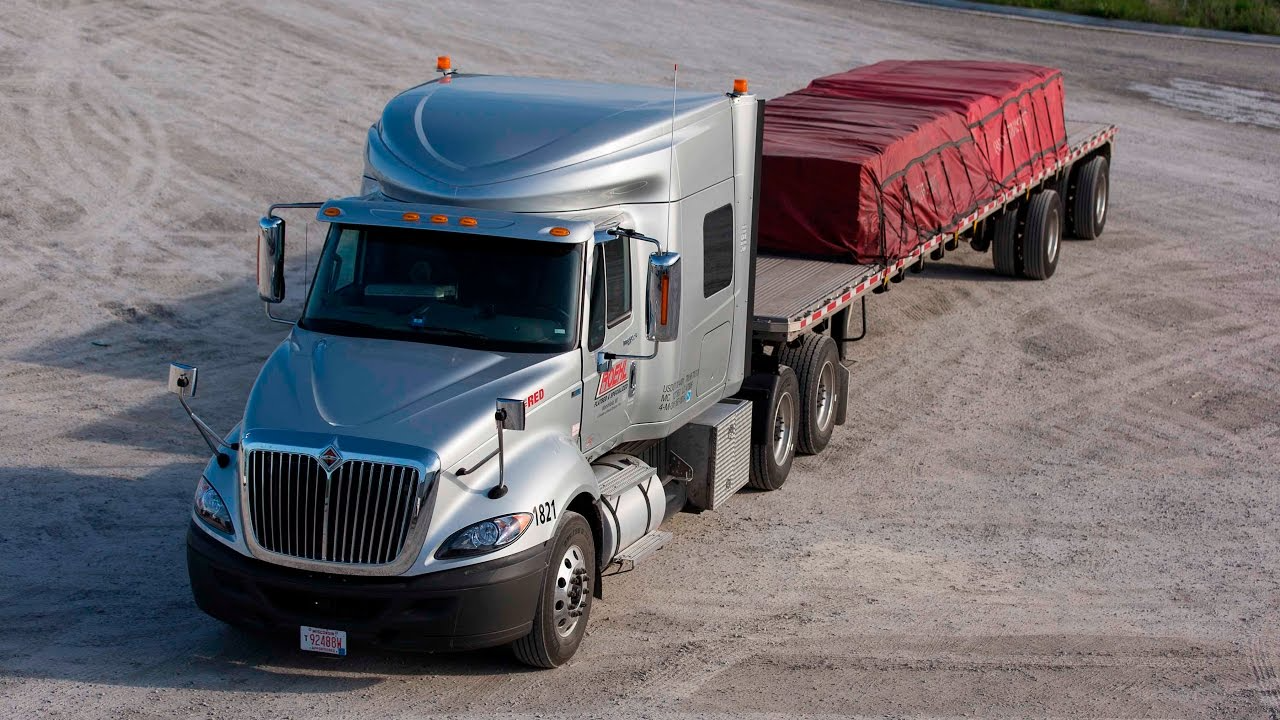A silver semi-truck with a flatbed trailer carries red cargo, parked on a gravel surface under a clear sky, representing flatbed truck rental Dubai services and suggesting transportation or logistics.