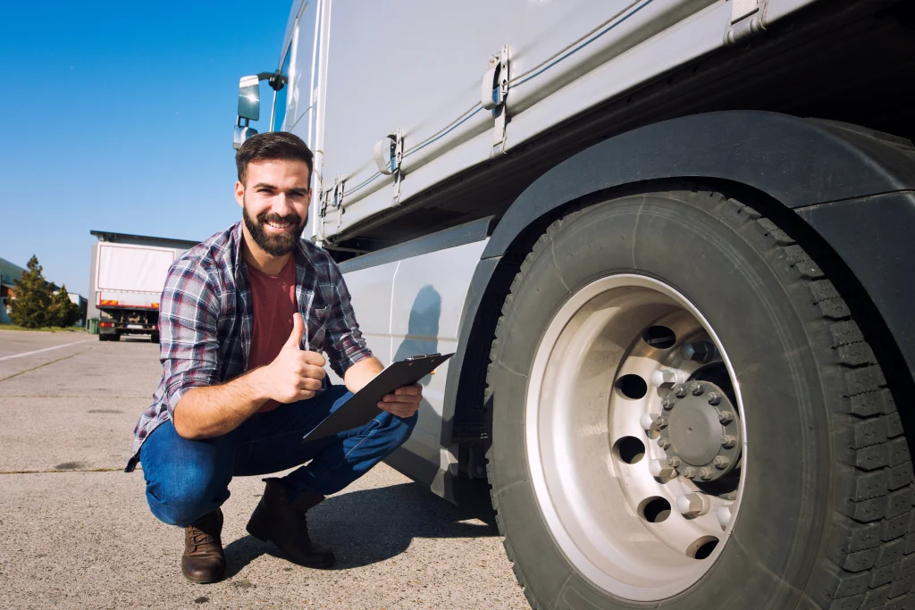 Man crouching by a truck's wheel giving a thumbs up on a sunny day, representing heavy truck rental Dubai services. He smiles, wearing a plaid shirt and jeans, conveying a positive and confident mood.