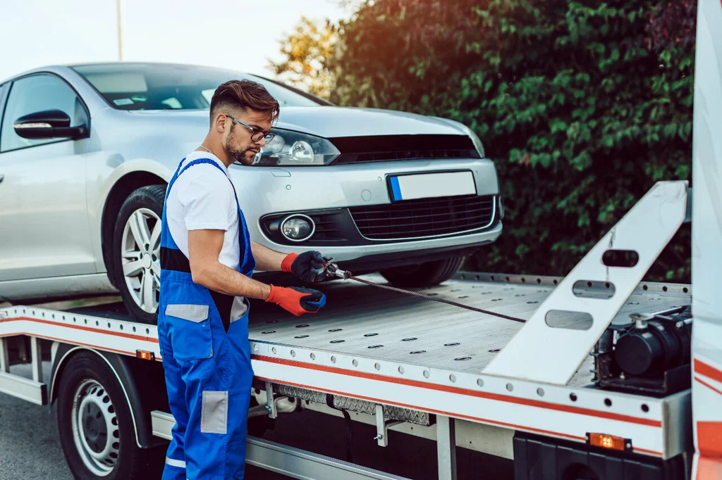 A tow truck operator in blue overalls secures a silver car onto a flatbed in daylight. The scene conveys efficiency and focus, with trees in the background.