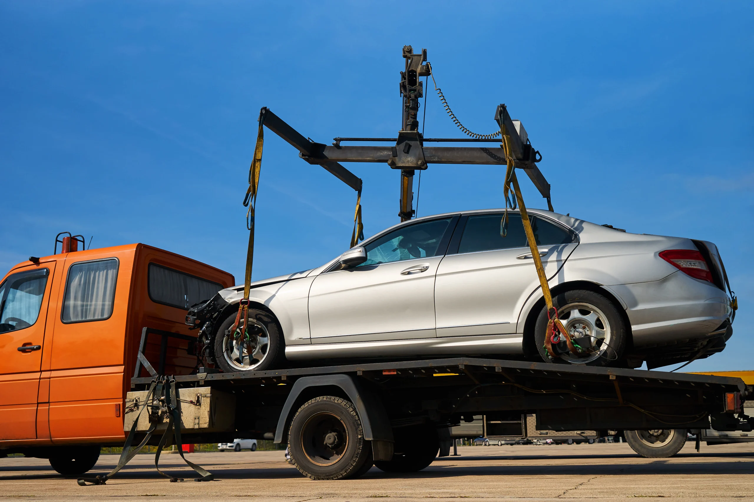 A silver car is secured on a flatbed tow truck under a clear blue sky. The orange truck contrasts with the car, conveying a tone of efficiency and resolve.