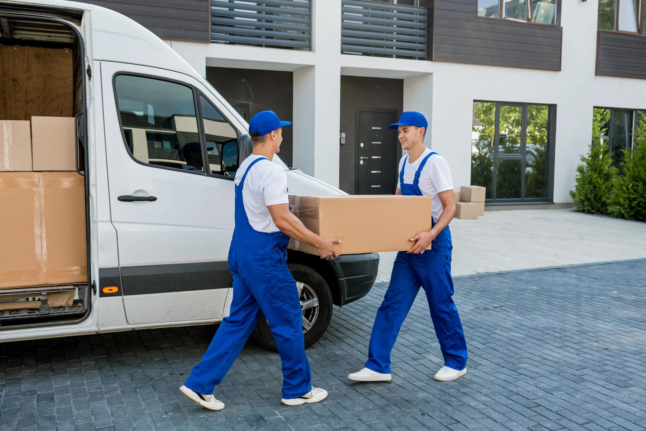 Two workers in blue uniforms carry a large cardboard box from a delivery van parked outside a modern building, representing pickup truck delivery Dubai services and conveying teamwork and efficiency.