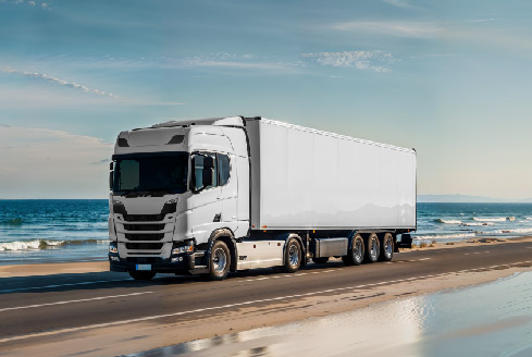 A white delivery truck drives along a coastal road under a clear blue sky, with the ocean and sandy beach in the background, showcasing reliable heavy truck rental Dubai.