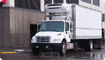 A white refrigerated truck is parked beside an industrial building. The vehicle is positioned near a loading bay on a wet street, implying a recent delivery.