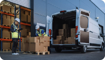 Two workers in safety gear load boxes onto a pallet next to an open delivery van at a warehouse, conveying teamwork and efficiency.