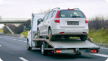 A white tow truck carries a silver car on a highway under a cloudy sky, evoking a sense of travel and urgency. An overpass is visible in the background.