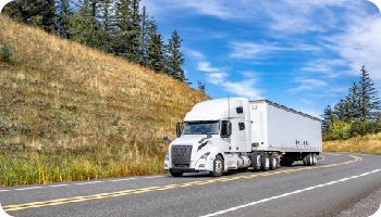 A white semi-truck drives down a scenic highway, bordered by grassy hills and pine trees, under a bright blue sky with scattered clouds.