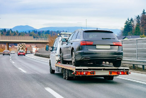 On a highway with mountains and trees, a tow truck carrying a black SUV highlights professional car recovery Dubai.