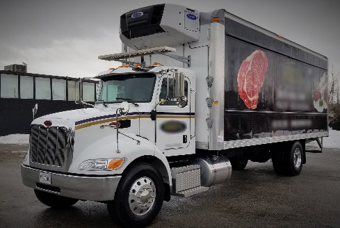 A white refrigerated truck is parked on a snowy day, with a graphic of raw meat on its side panel, overcast sky and industrial buildings in the background, showcasing professional chiller truck rental Dubai services.