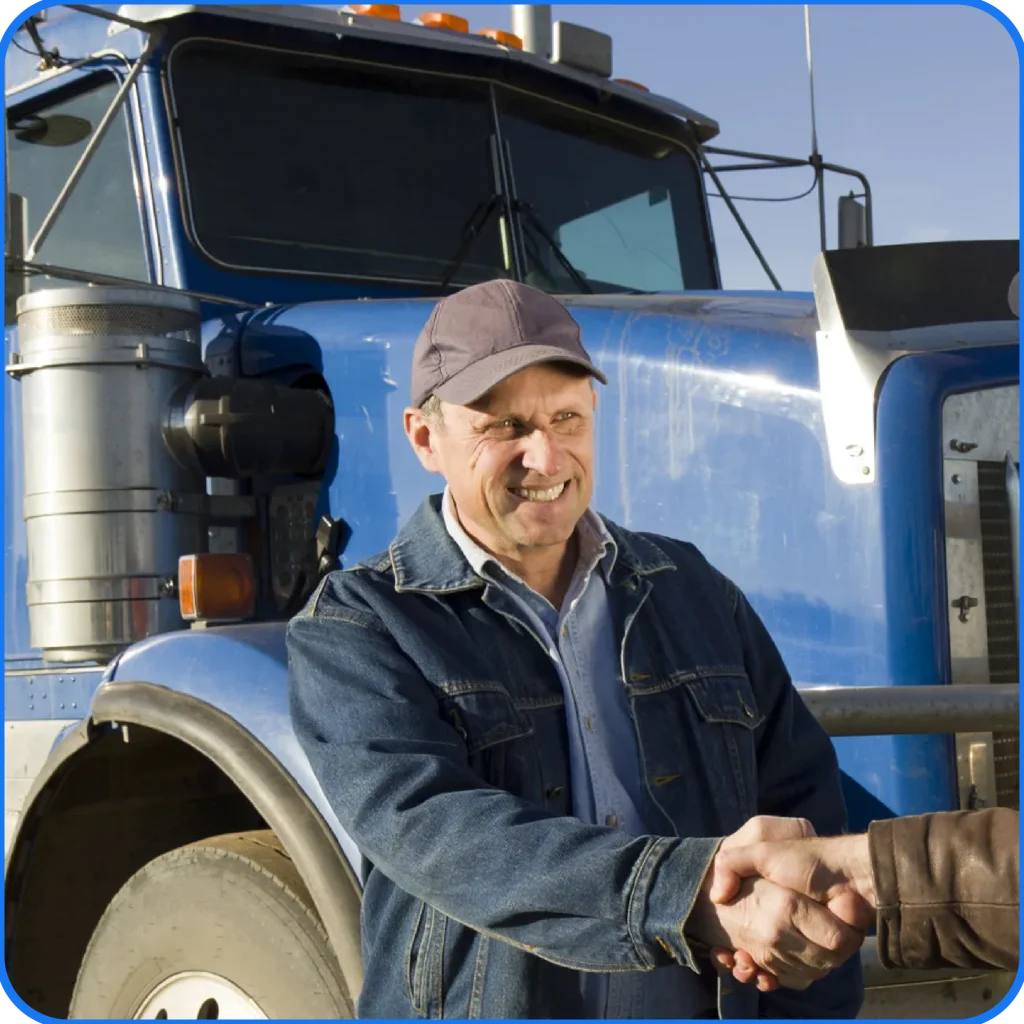 Smiling man in a denim jacket and cap shakes hands in front of a large blue truck under a clear sky, conveying friendliness and agreement.