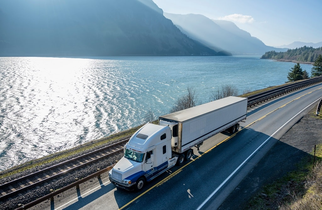 On a scenic lakeside road with mountains, a white semi-truck highlights professional chiller truck rental Dubai for safe and efficient transport.
