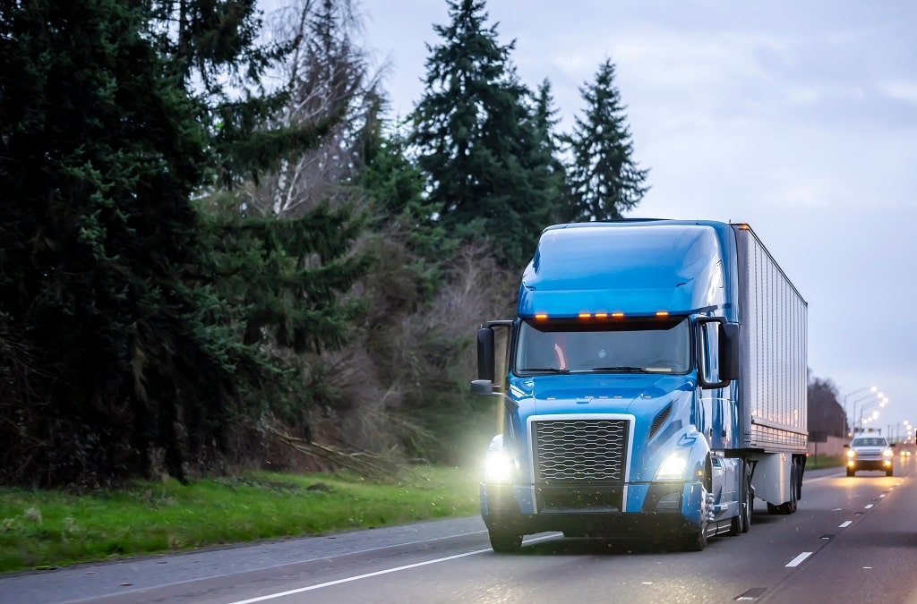 A blue semi-truck with headlights on drives along a rural road lined with tall evergreen trees under a cloudy sky, showcasing reliable chiller truck rental Dubai services.