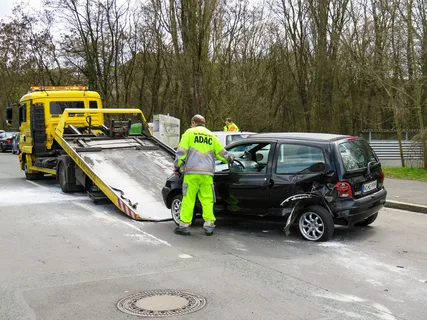 Tow truck loading a damaged black car after an accident on a wooded roadside, with a worker in a bright yellow and gray uniform supervising — part of Drivelo Services.